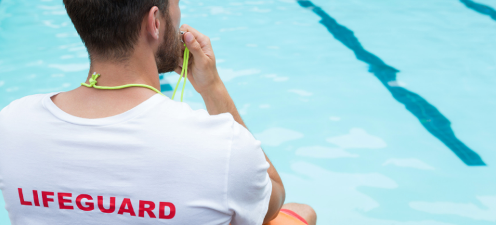 Man wearing lideguard tshirt sitting by pool, blowing a whistle