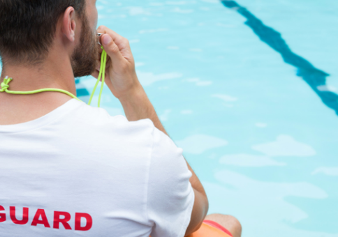Man wearing lifeguard t-shirt sitting by pool, blowing a whistle