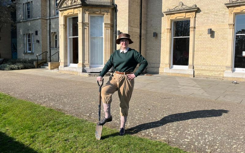 Visitor Host, Rachel, dressed as a Land Girl stood outside Chelmsford Museum with her foot on a spade.