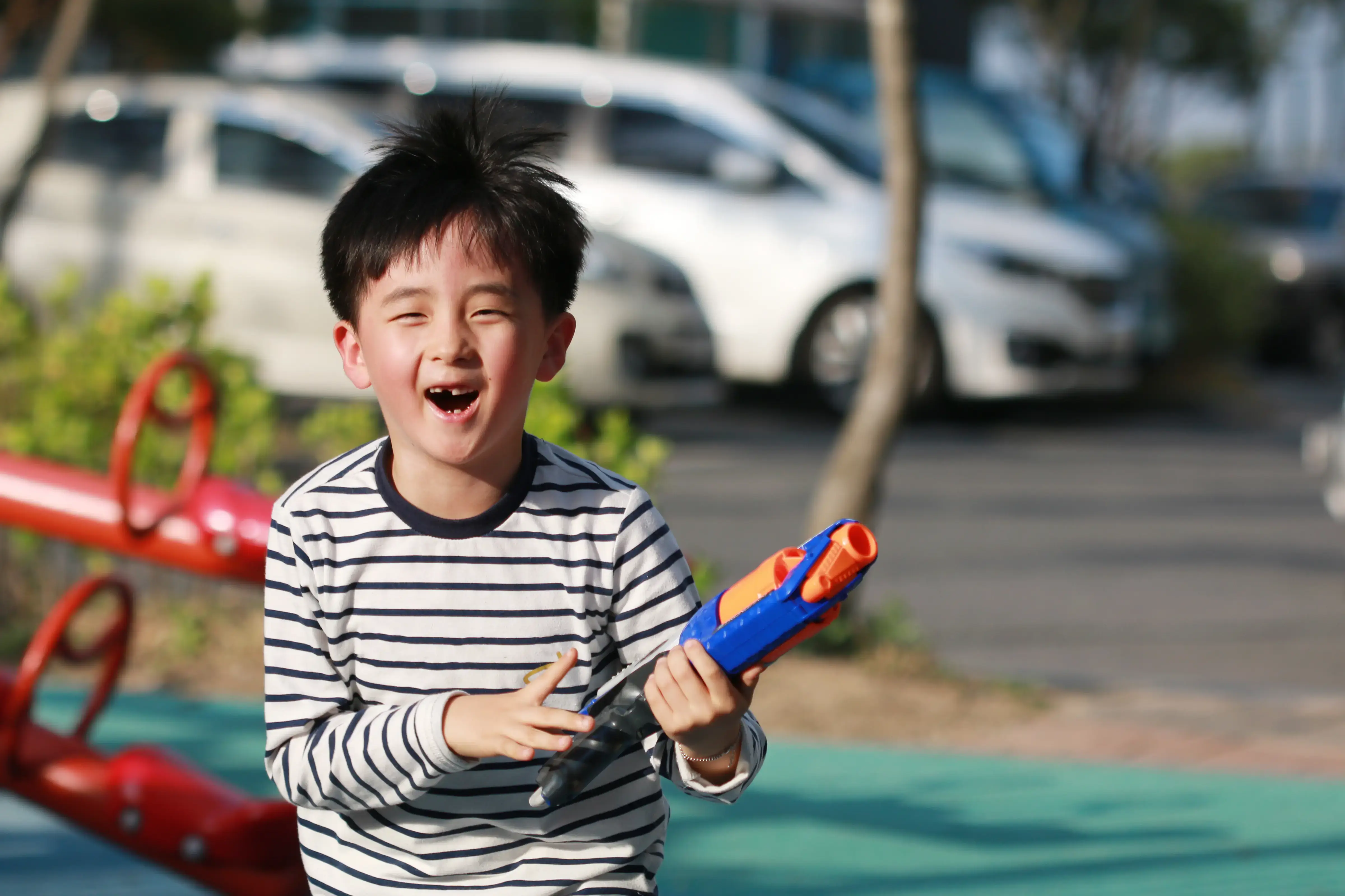 Young boy with missing tooth wearing white top with black stripes, smiling and holding a blue and orange Nerf gun