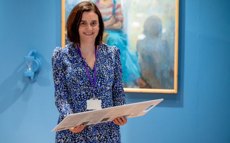 A woman with shoulder length brown hair and a patterned blue dress smiles at the camera while standing in the Shifting Perspectives exhibition. 