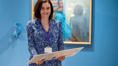 A woman with shoulder length brown hair and a patterned blue dress smiles at the camera while standing in the Shifting Perspectives exhibition. 
