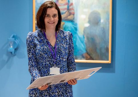 A woman with shoulder length brown hair and a patterned blue dress smiles at the camera while standing in the Shifting Perspectives exhibition. 