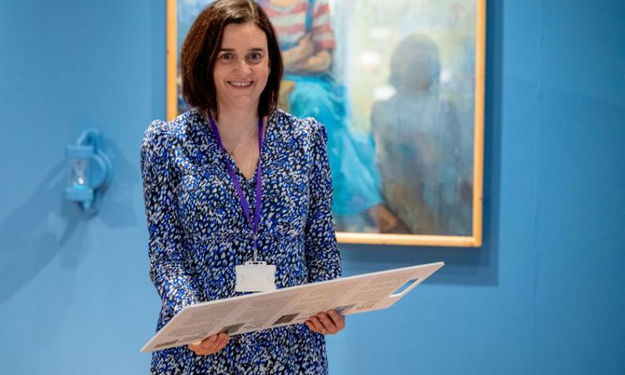 A woman with shoulder length brown hair and a patterned blue dress smiles at the camera while standing in the Shifting Perspectives exhibition. 