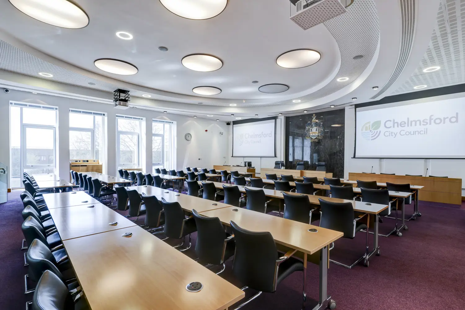 Large, well-lit room with rows of long tables and chairs laid out facing big screens