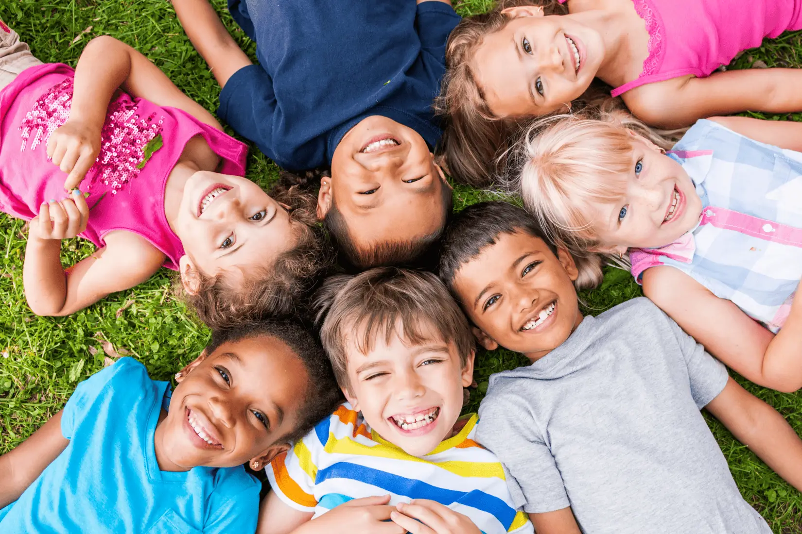 Group of happy young children lying on the grass in the sunshine a circle with their heads in the middle.