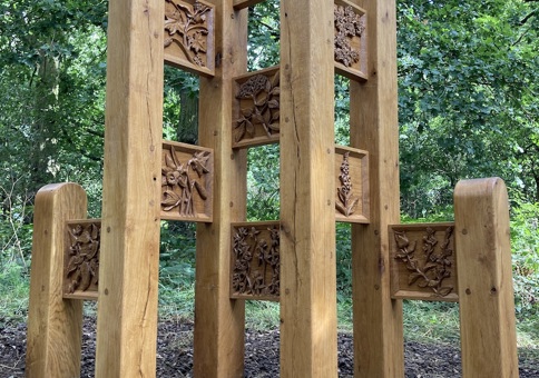 Large oak sculpture featuring tall posts and carved square panels featuring reliefs of flowers and other flora