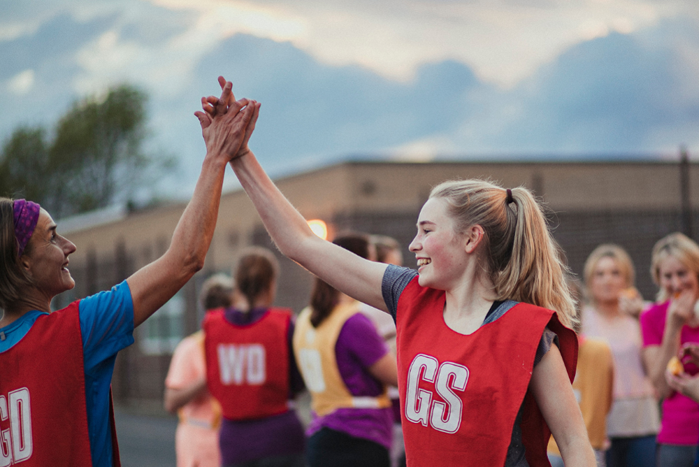 Two netball players, one older and one younger, high-fiving with other team members in the background.
