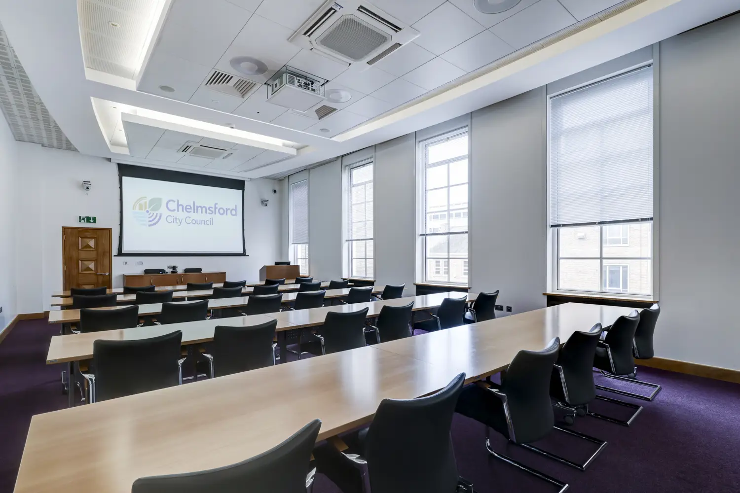 Meeting room set out classroom style, with tables and chairs in long rows facing large screen