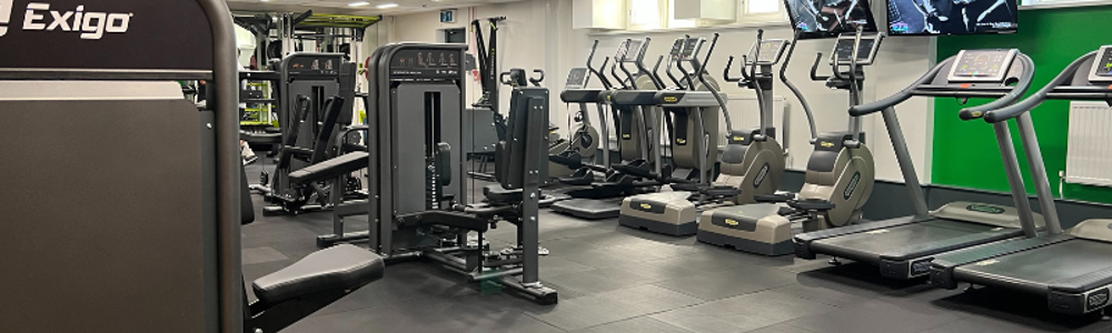A row of treadmills, cross trainers, and cycle machines against a wall in a gym, and other cardiovascular machines in the centre.