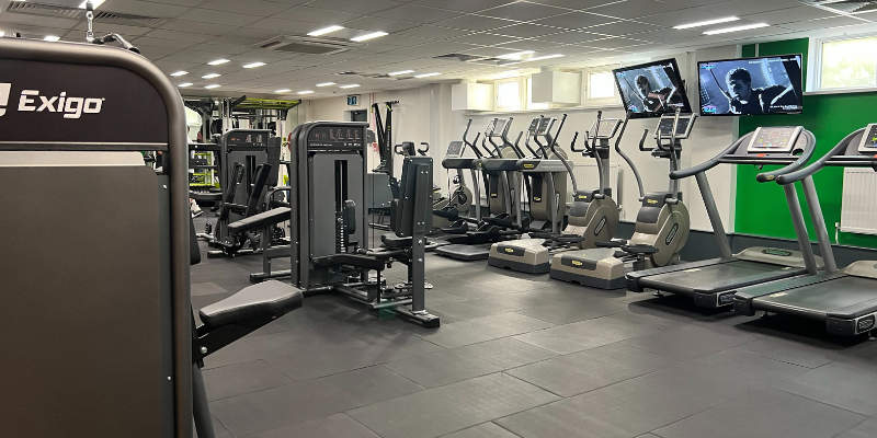 A row of treadmills, cross trainers, and cycle machines against a wall in a gym, and other cardiovascular machines in the centre.
