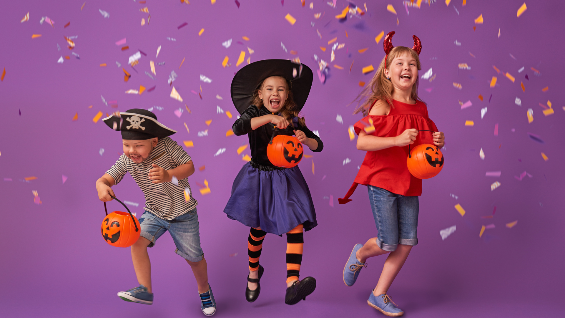 Kids in Halloween fancy dress jumping and smiling in front of confetti and a purple background.