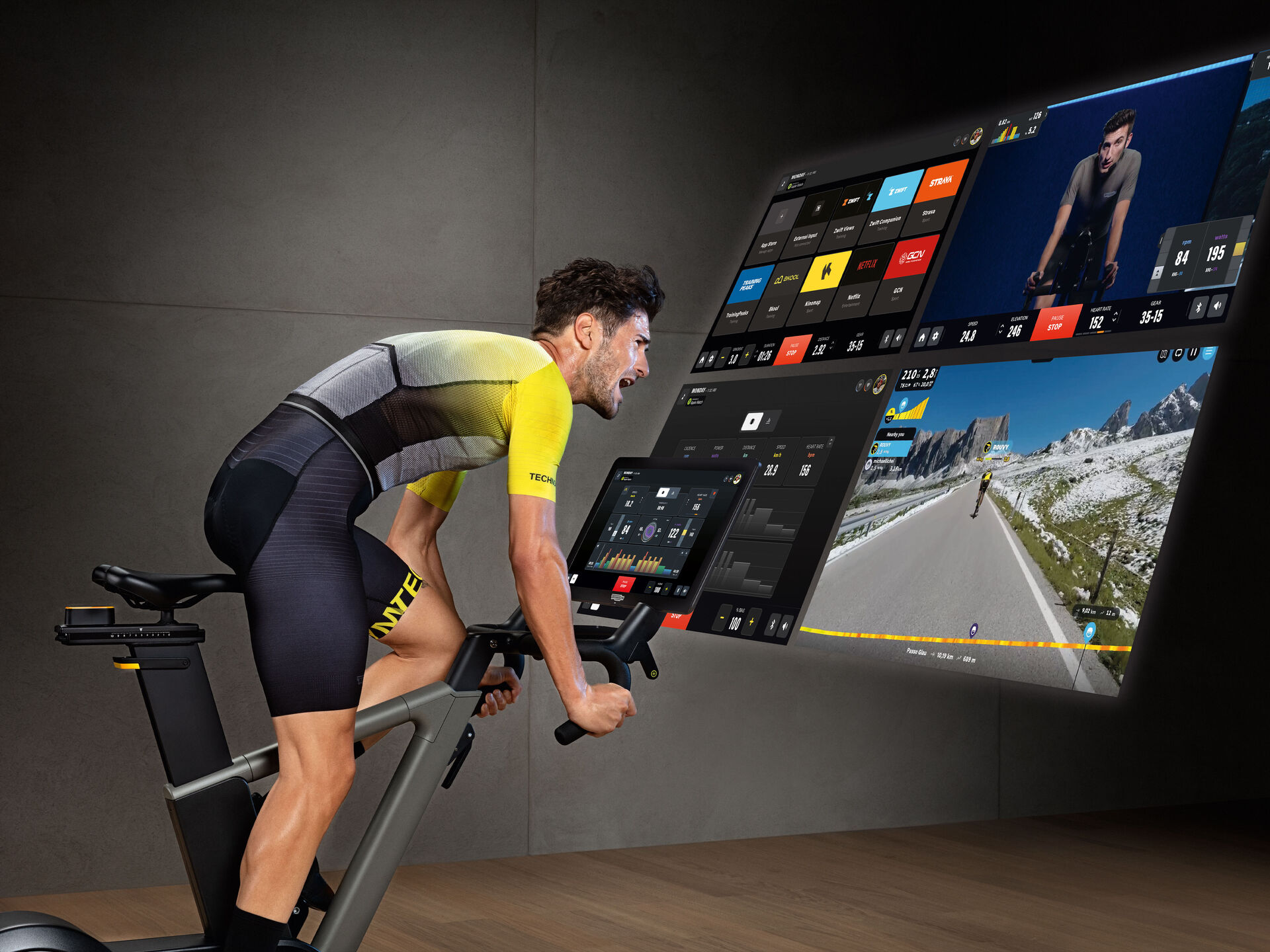 Side image of a man in lycra riding a stationary bike in a gym, while watching a large digital screen displaying a virtual route.