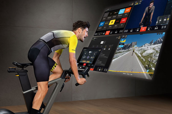 Side image of a man in lycra riding a stationary bike in a gym, while watching a large digital screen displaying a virtual route.