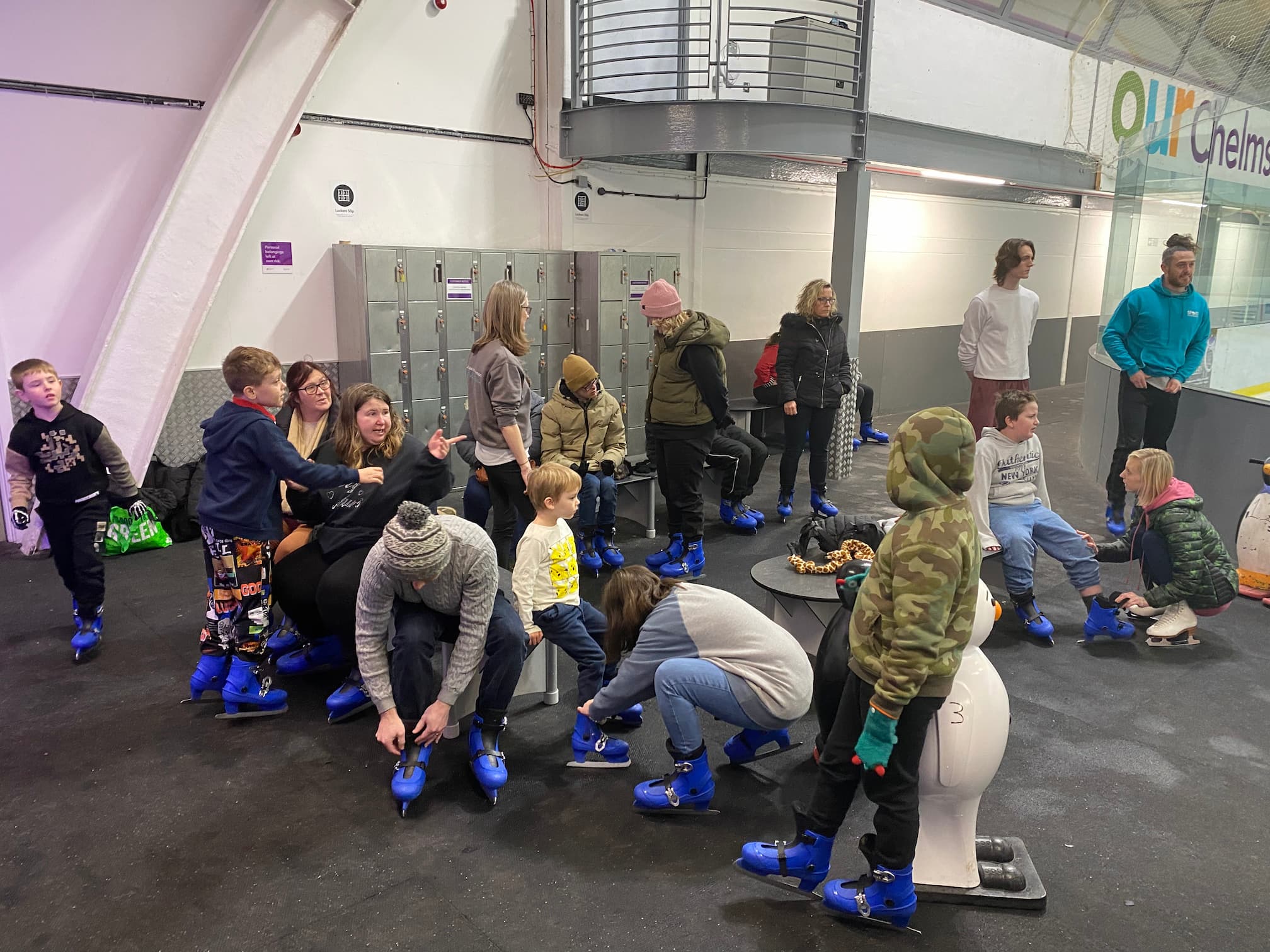 Group of adults and children sat on benches putting on blue ice skates at an indoor ice rink