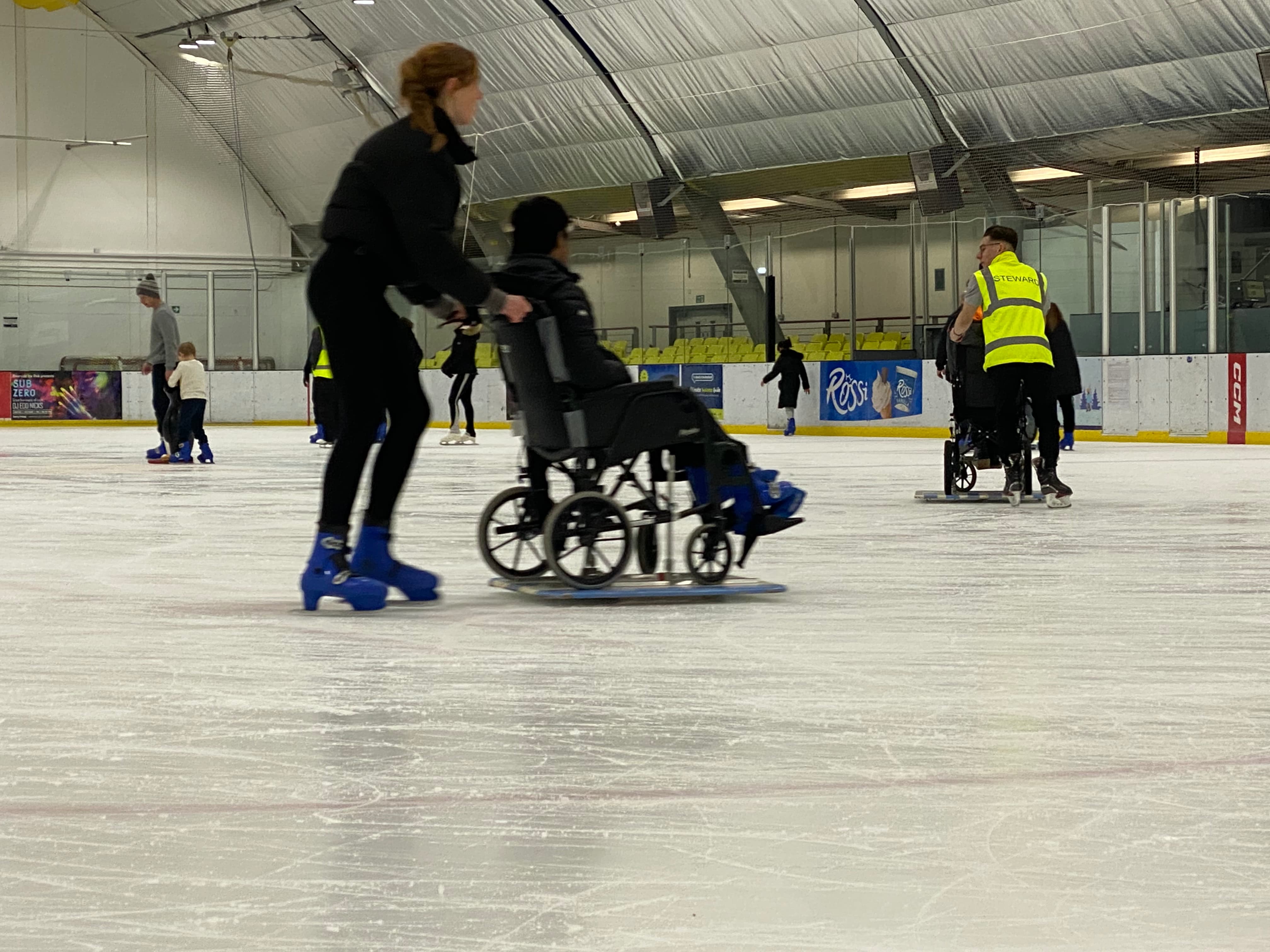 Young woman on ice skates in an ice rink, pushing a person in a wheelchair along on the ice