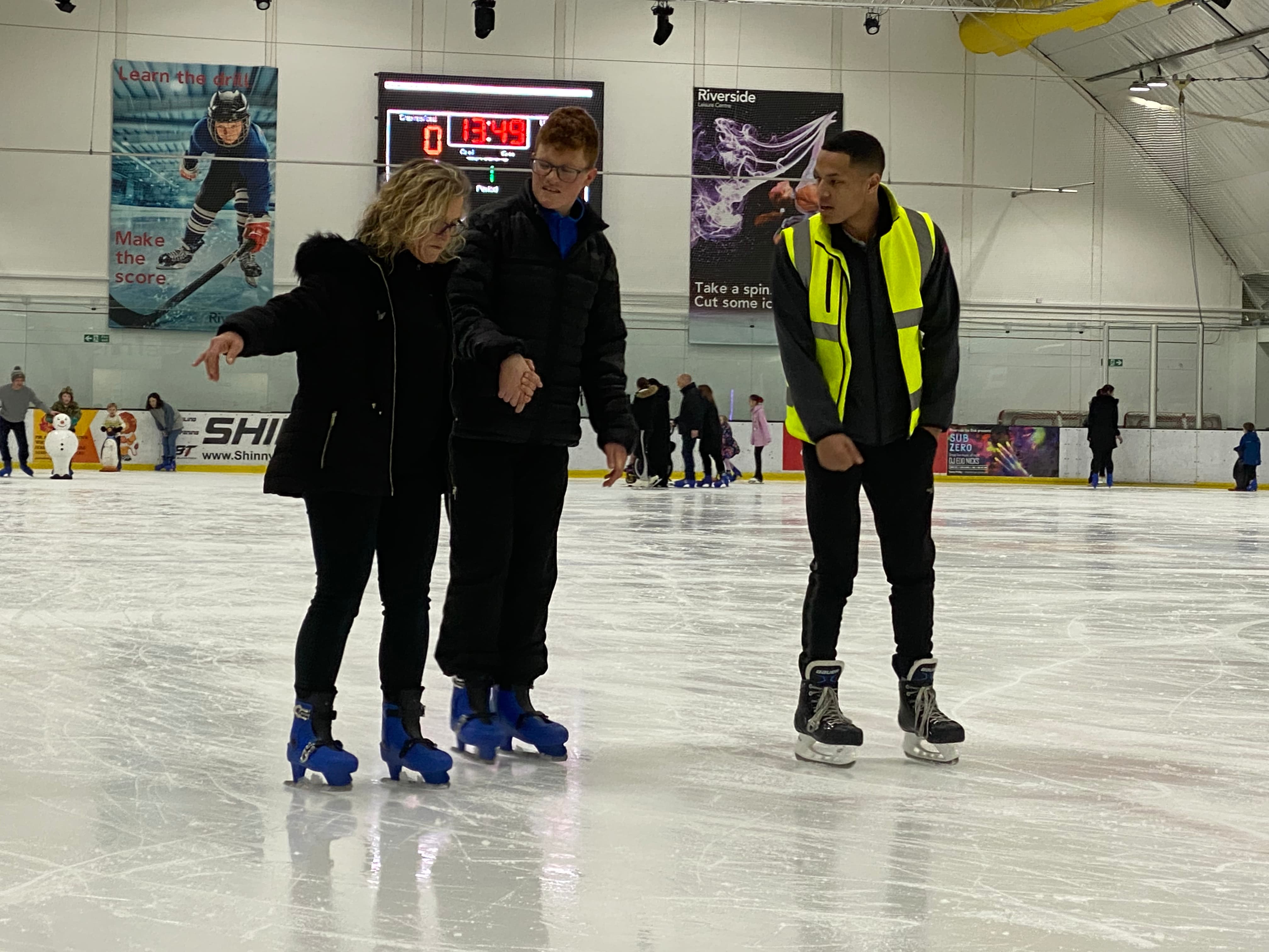 Two people learning to skate, being taught by a man in a high visibility jacket