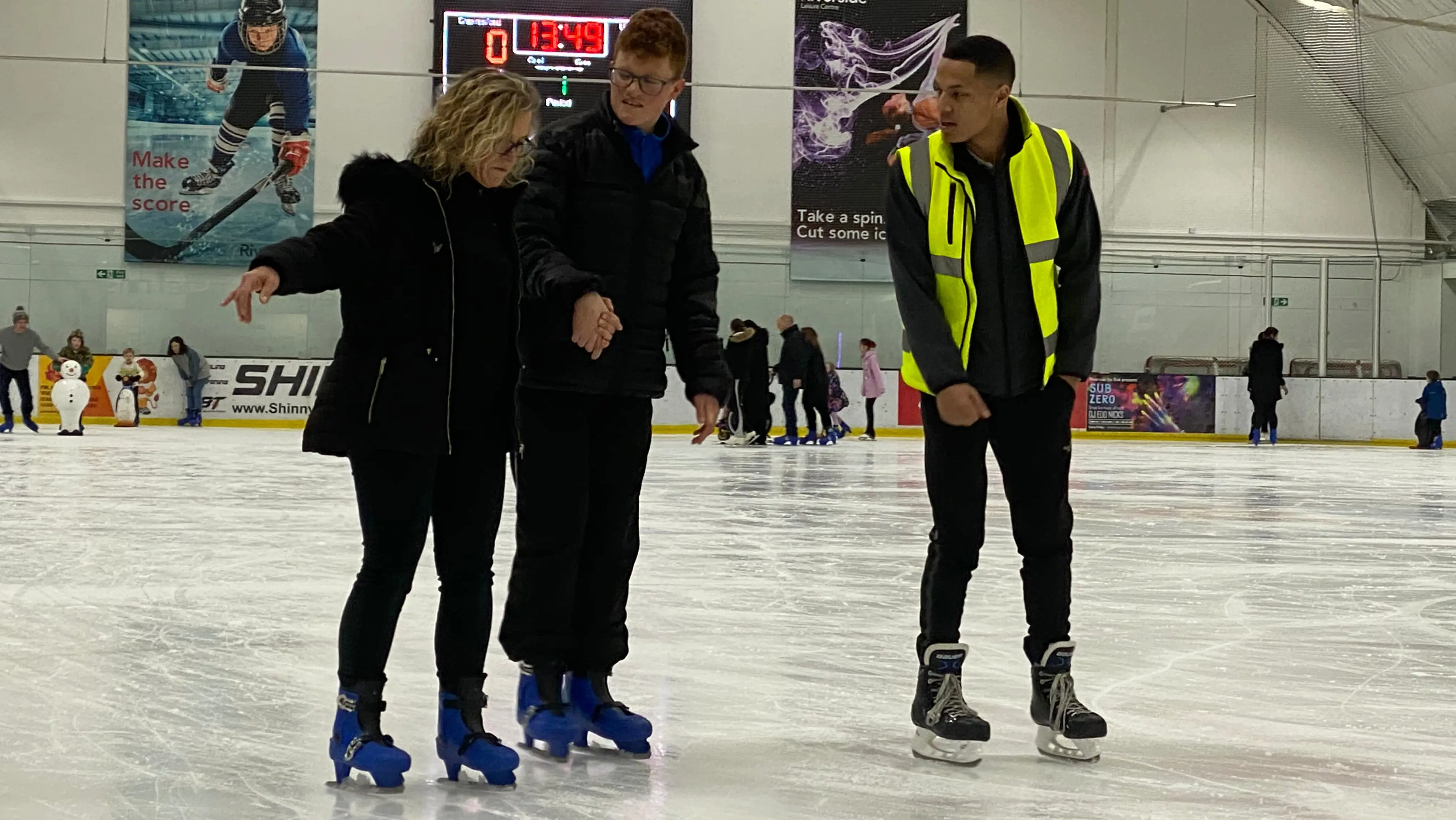 Two people learning to skate, being taught by a man in a high visibility jacket