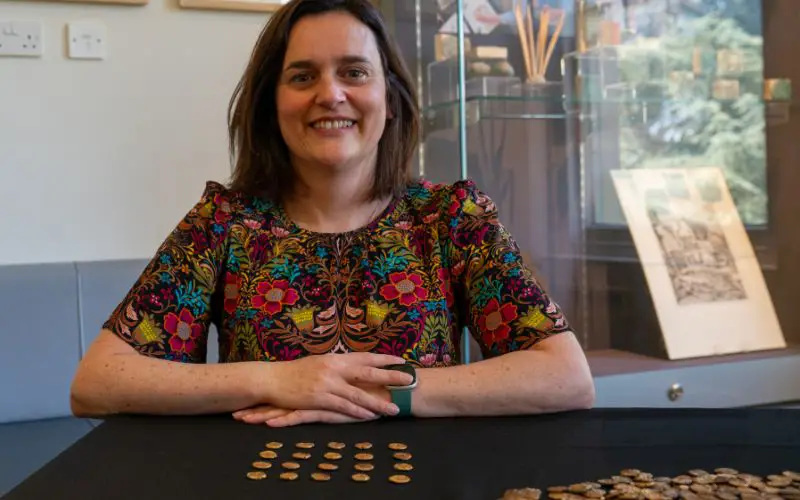 Curator Claire Willets smiles while sat by a table filled with gold coins. 