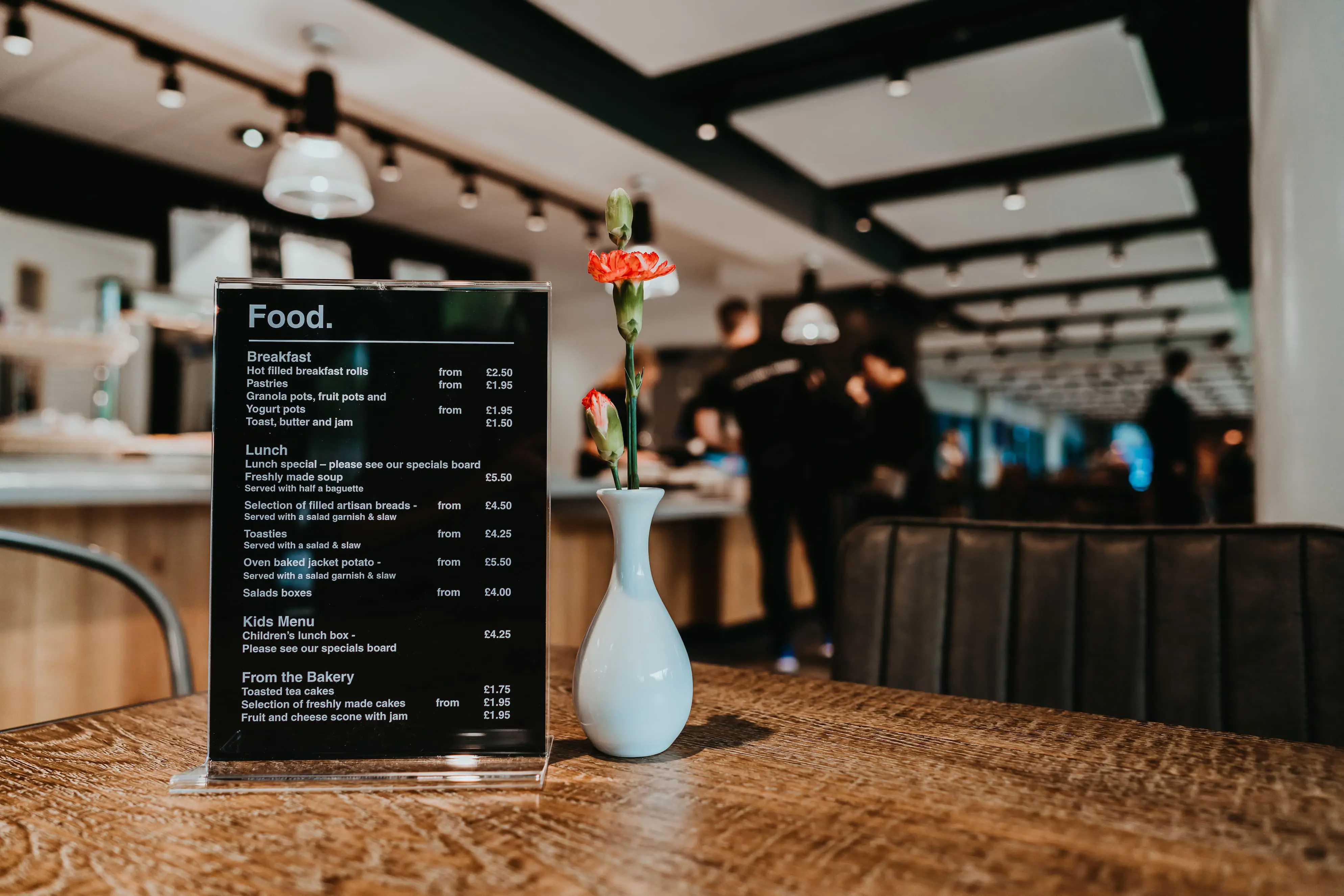 Small food menu sitting on wooden table with small white vase containing carnations