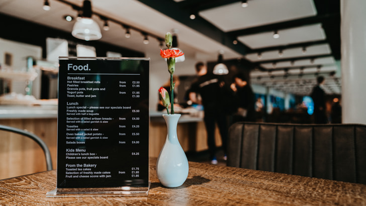 Small food menu sitting on wooden table with small white vase containing carnations