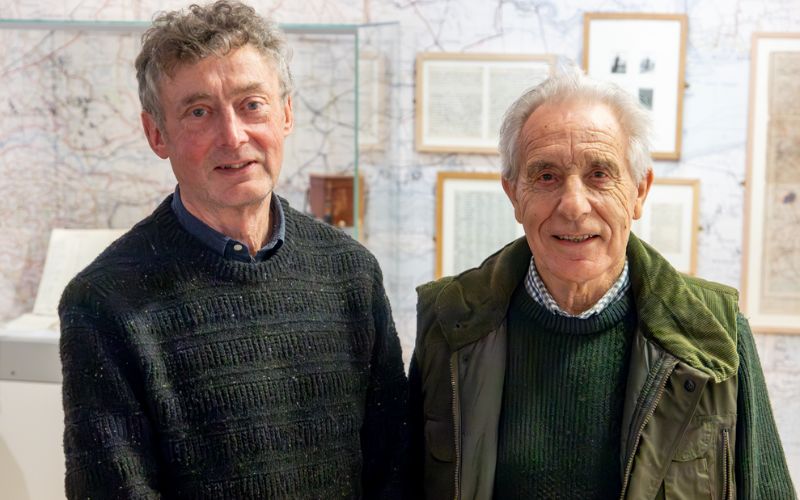 Two men in jumpers smiling at the camera. Behind them are maps on the wall and diaries of J.A. Baker on display. 