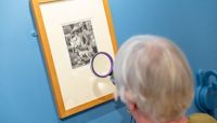 A woman looks closely at a piece of art with a magnifying glass.  preview