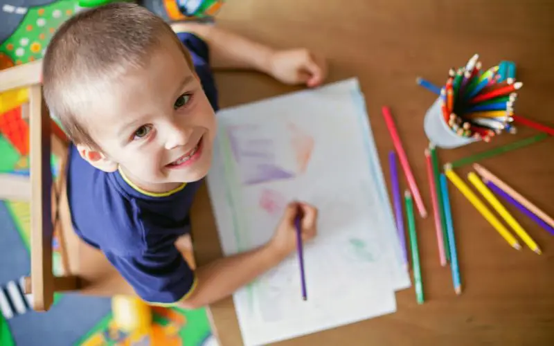 A boy smiles up at the camera while drawing with coloured pencils. 