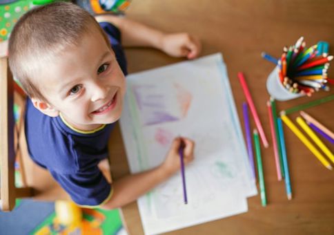 A boy smiles up at the camera while drawing with coloured pencils. 