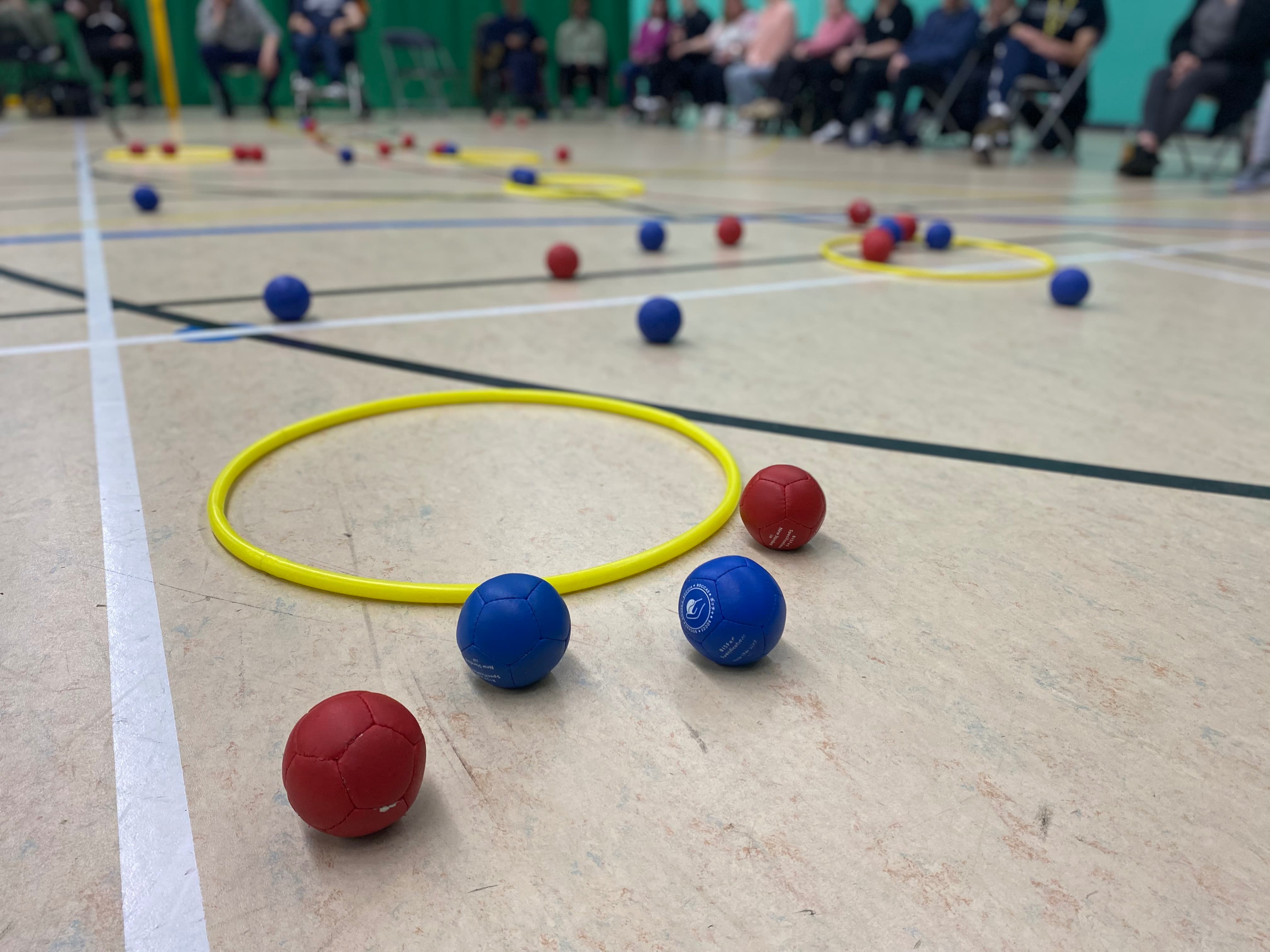 red and blue Boccia balls on the floor of a sports hall, next to a yellow hoop