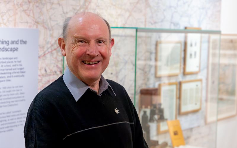 David Simmonds smiling inside the J.A. Baker exhibition. Behind him is a glass case filled with Baker's items, such as binoculars and journals. 