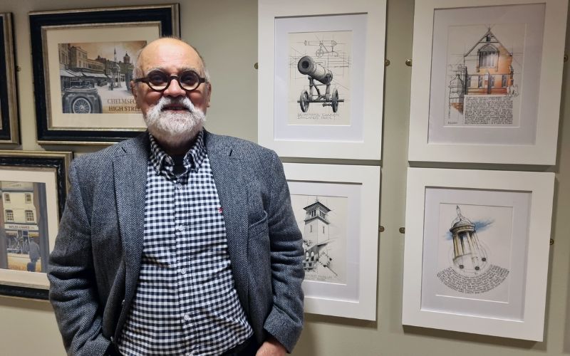 An older man with grey hair and dark circular glasses stands in front of a wall with various framed artwork hung. 
