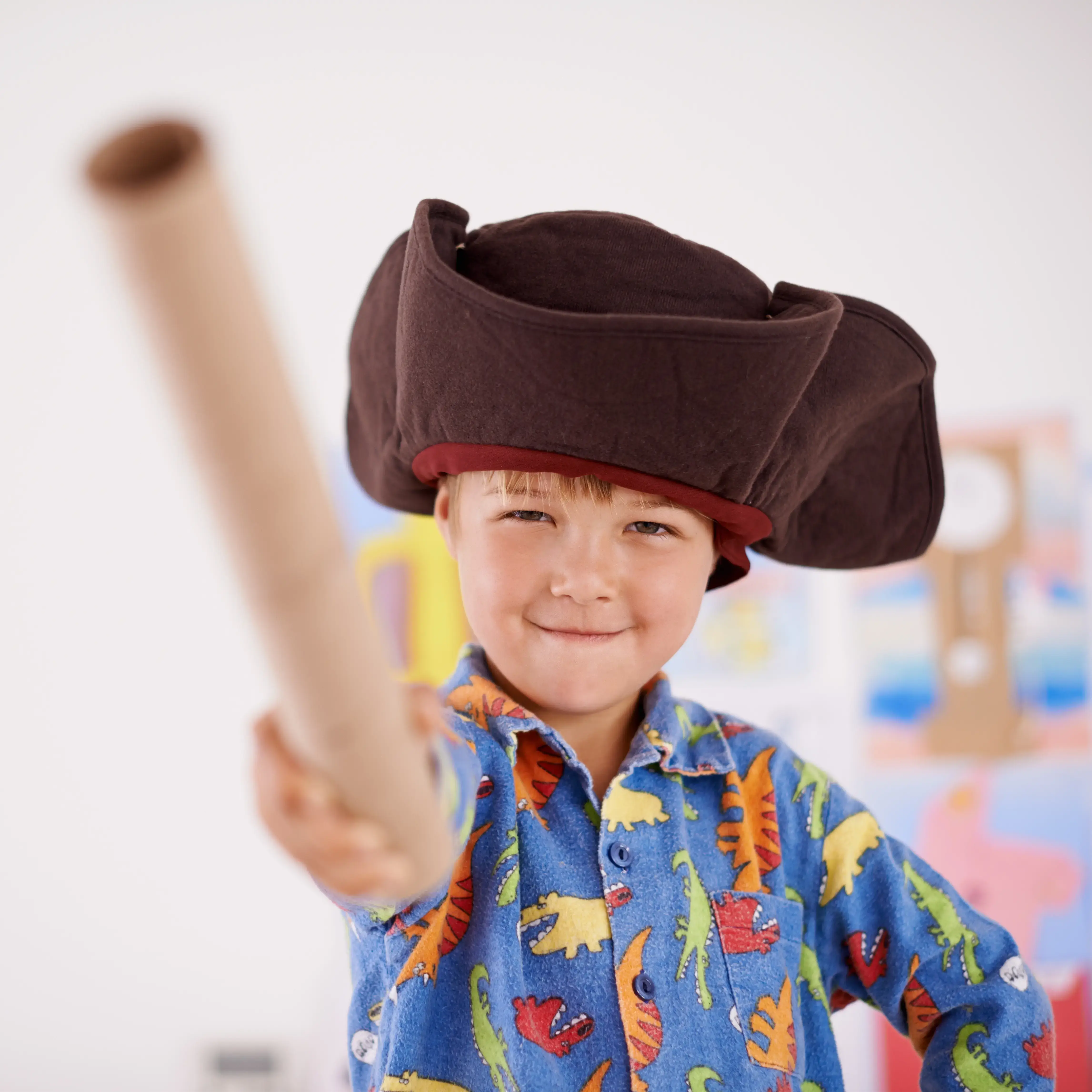 Boy wearing blue short with dinosaurs on it, with a black pirates hat and brandishing a cardboard tube like a sword