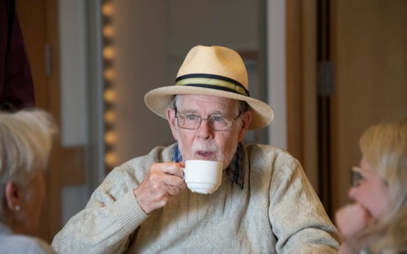A man sipping a cup of tea while sat around a table with others.  preview