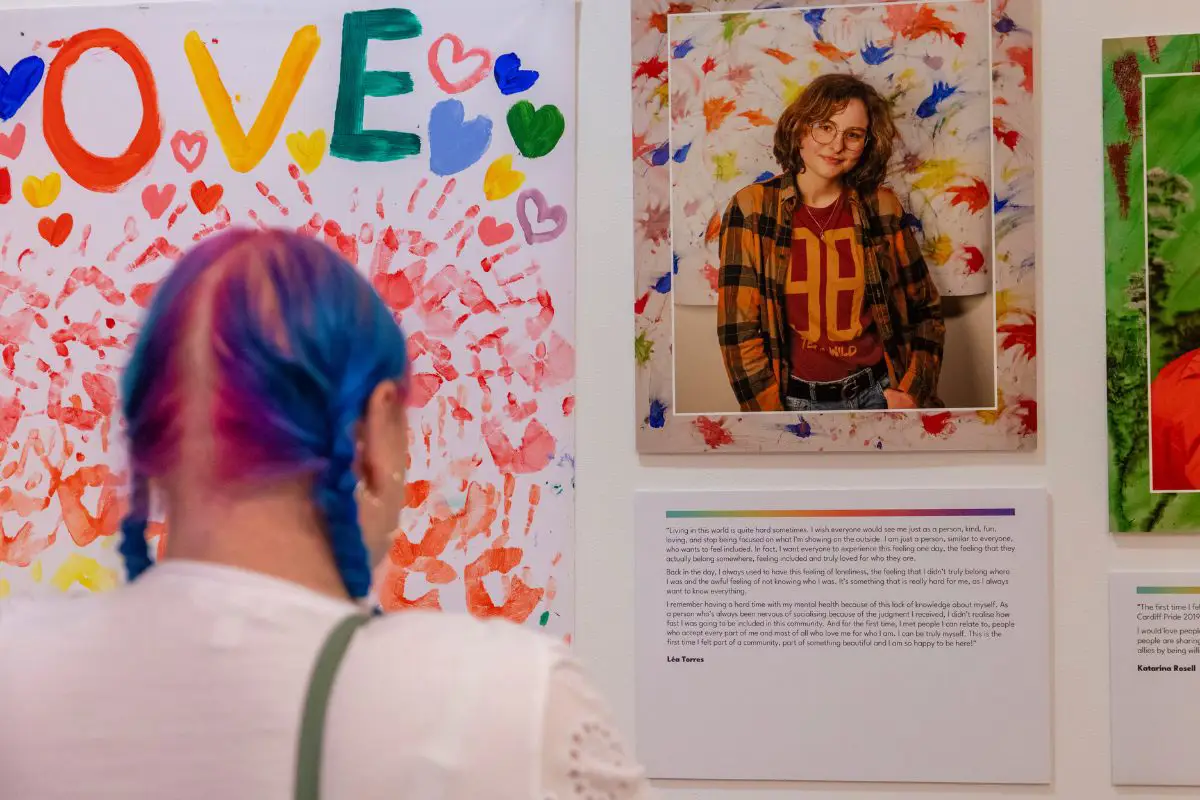 A person with bright blue coloured hair reads a poem displayed on the wall at the Behind the Rainbow exhibition. 