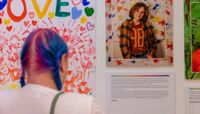 A person with bright blue coloured hair reads a poem displayed on the wall at the Behind the Rainbow exhibition.  preview