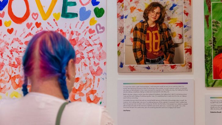 A person with bright blue coloured hair reads a poem displayed on the wall at the Behind the Rainbow exhibition. 