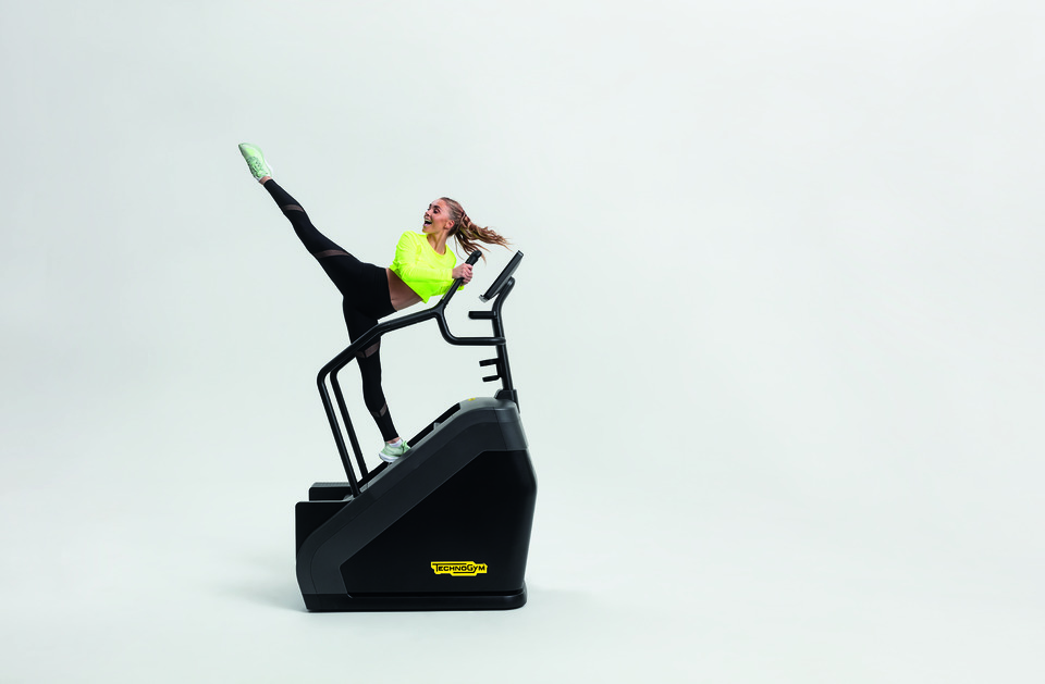 Energetic woman stood on a piece of stairclimber gym equipment, holding the handle and performing a high kick