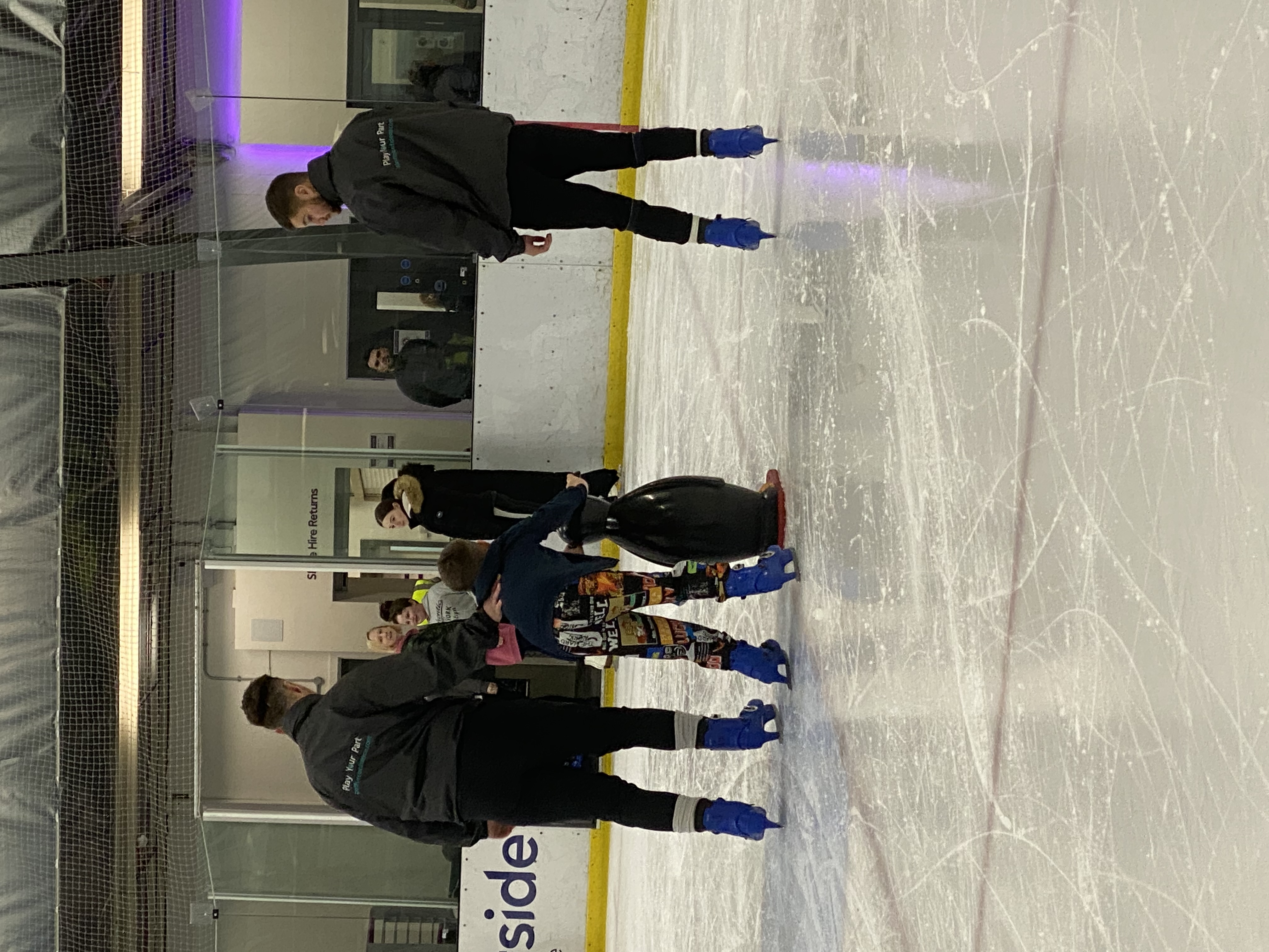 Young boy learning to ice skate, holding onto a penguin aid, with coaches either side.