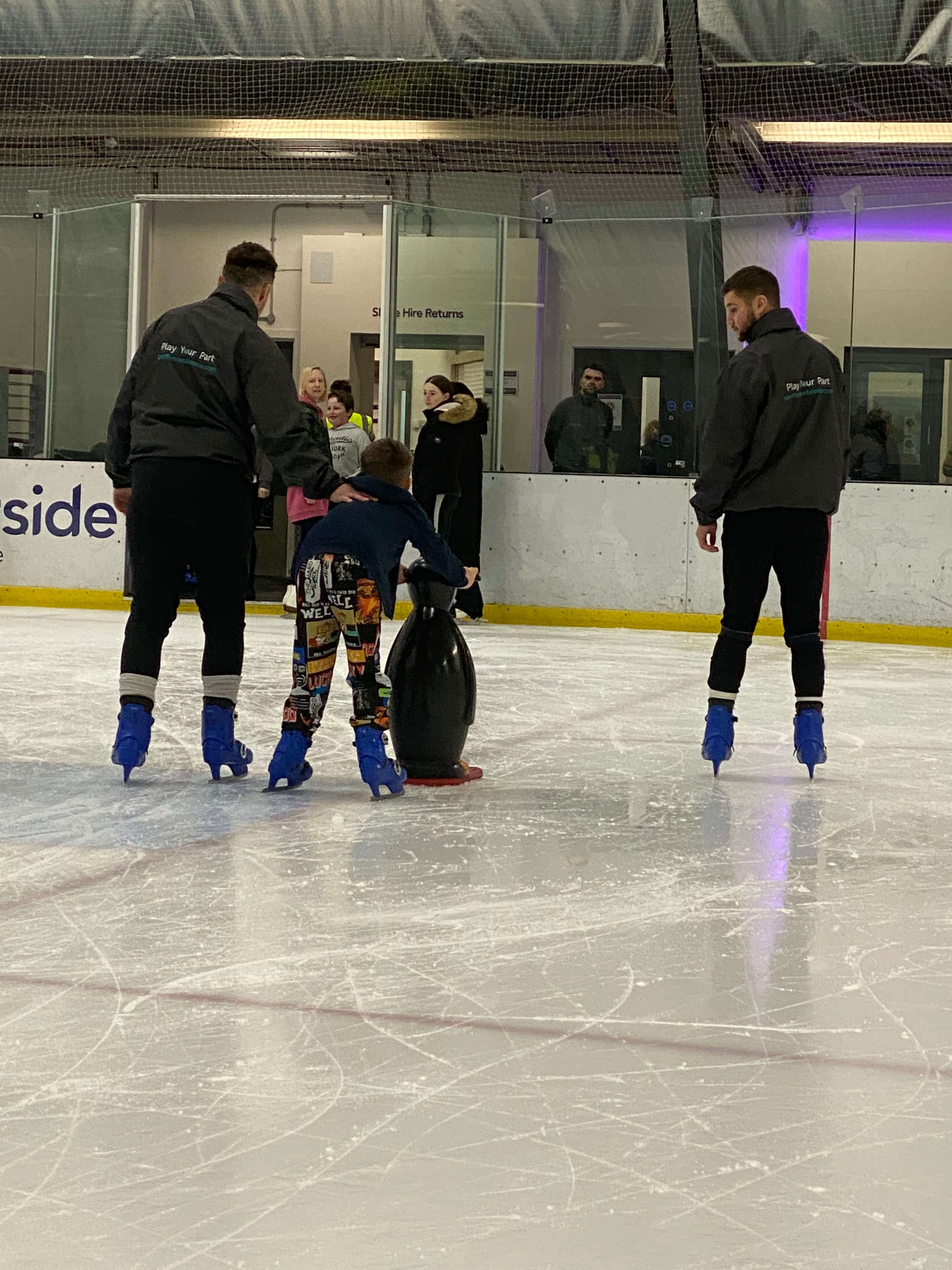Young boy learning to ice skate, holding onto a penguin aid, with coaches either side.