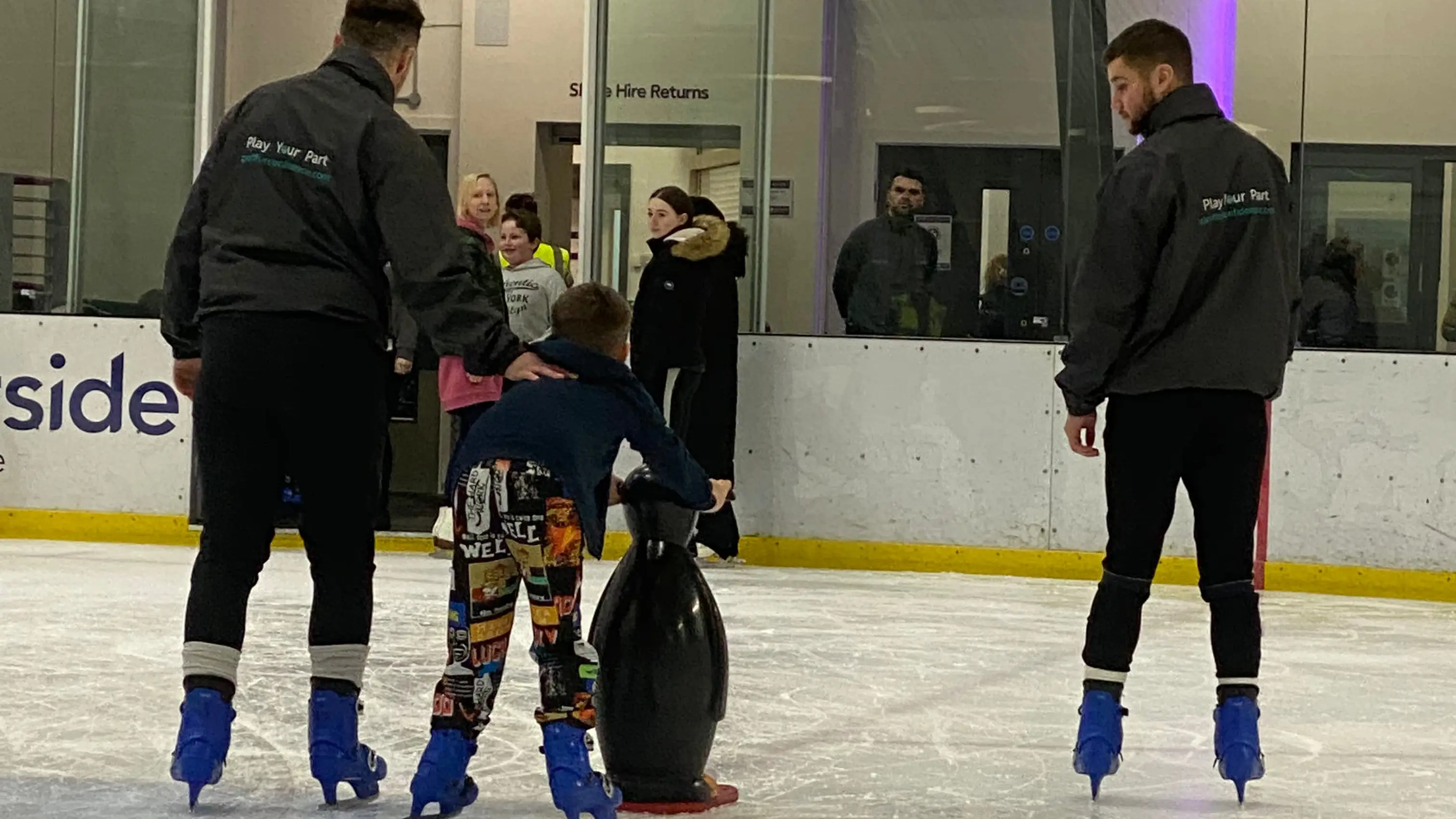 Young boy learning to ice skate, holding onto a penguin aid, with coaches either side.