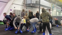 Group of adults and children sat on benches putting on blue ice skates at an indoor ice rink preview