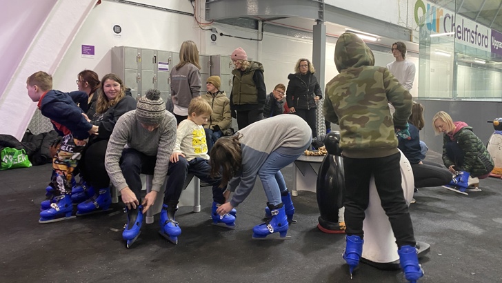 Group of adults and children sat on benches putting on blue ice skates at an indoor ice rink