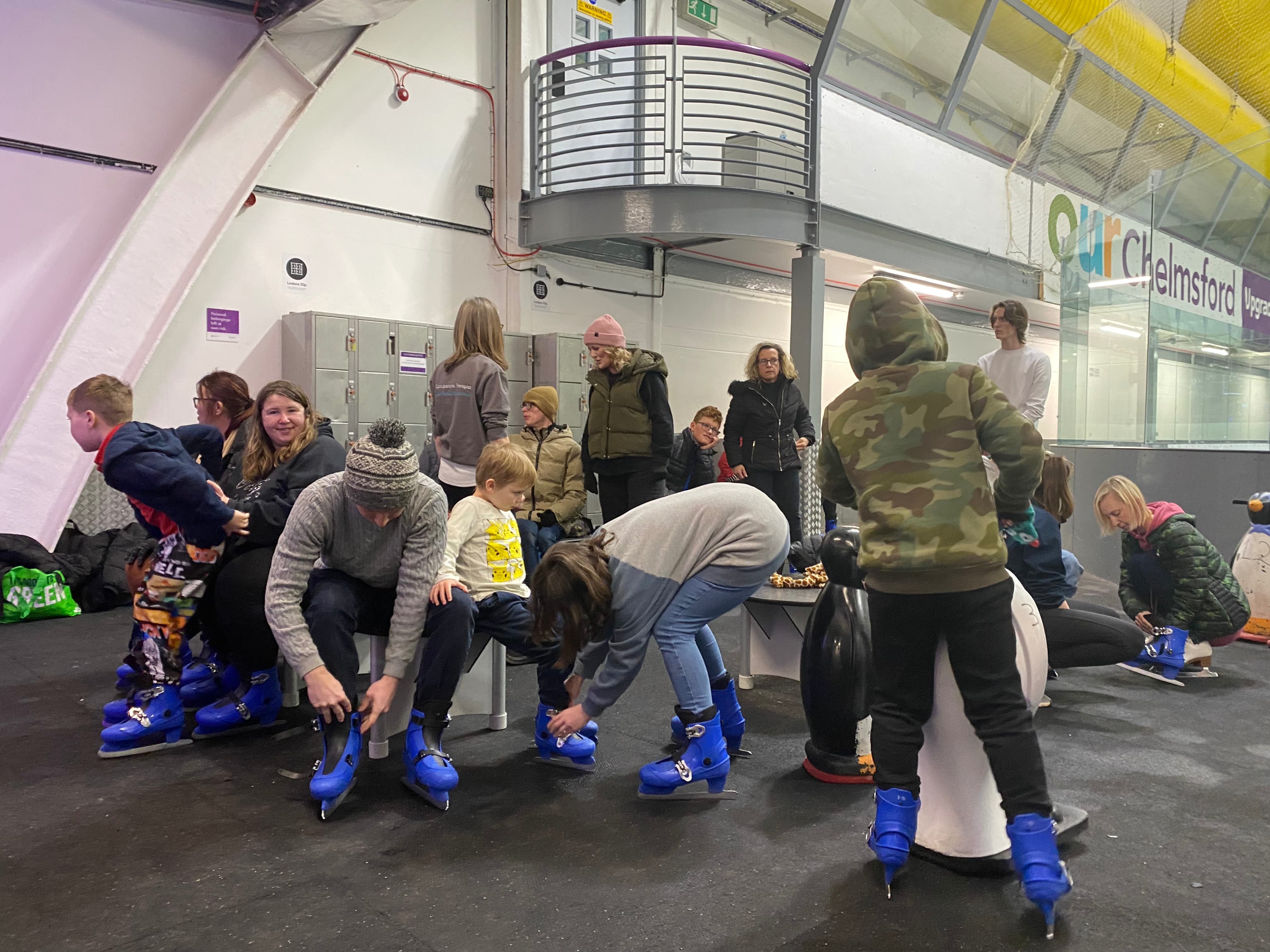 Group of adults and children sat on benches putting on blue ice skates at an indoor ice rink