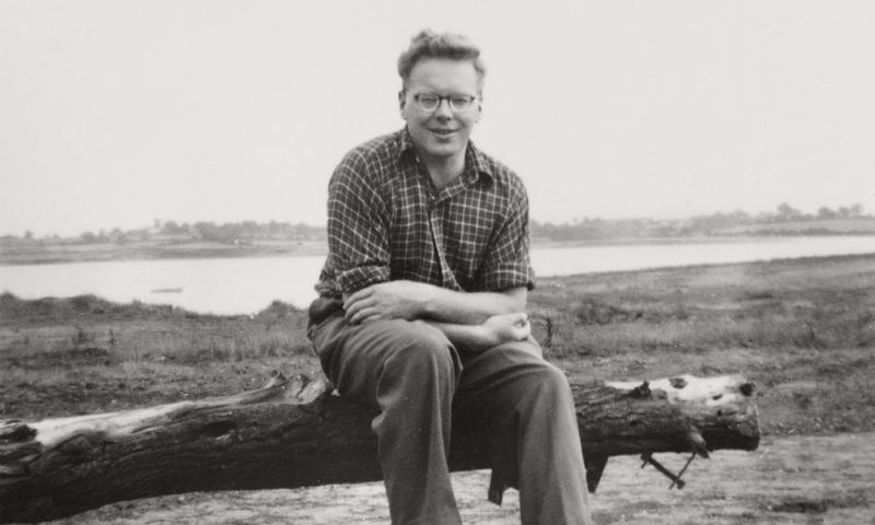 A black and white photo of JA Baker sitting on a log by the waterside. 