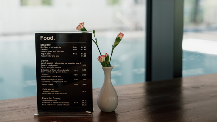 Small food menu sitting on wooden table with small white vase containing carnations, with a view of swimming pool behind