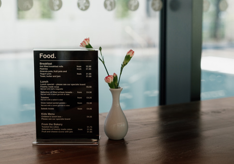 Small food menu sitting on wooden table with small white vase containing carnations, with a view of swimming pool behind