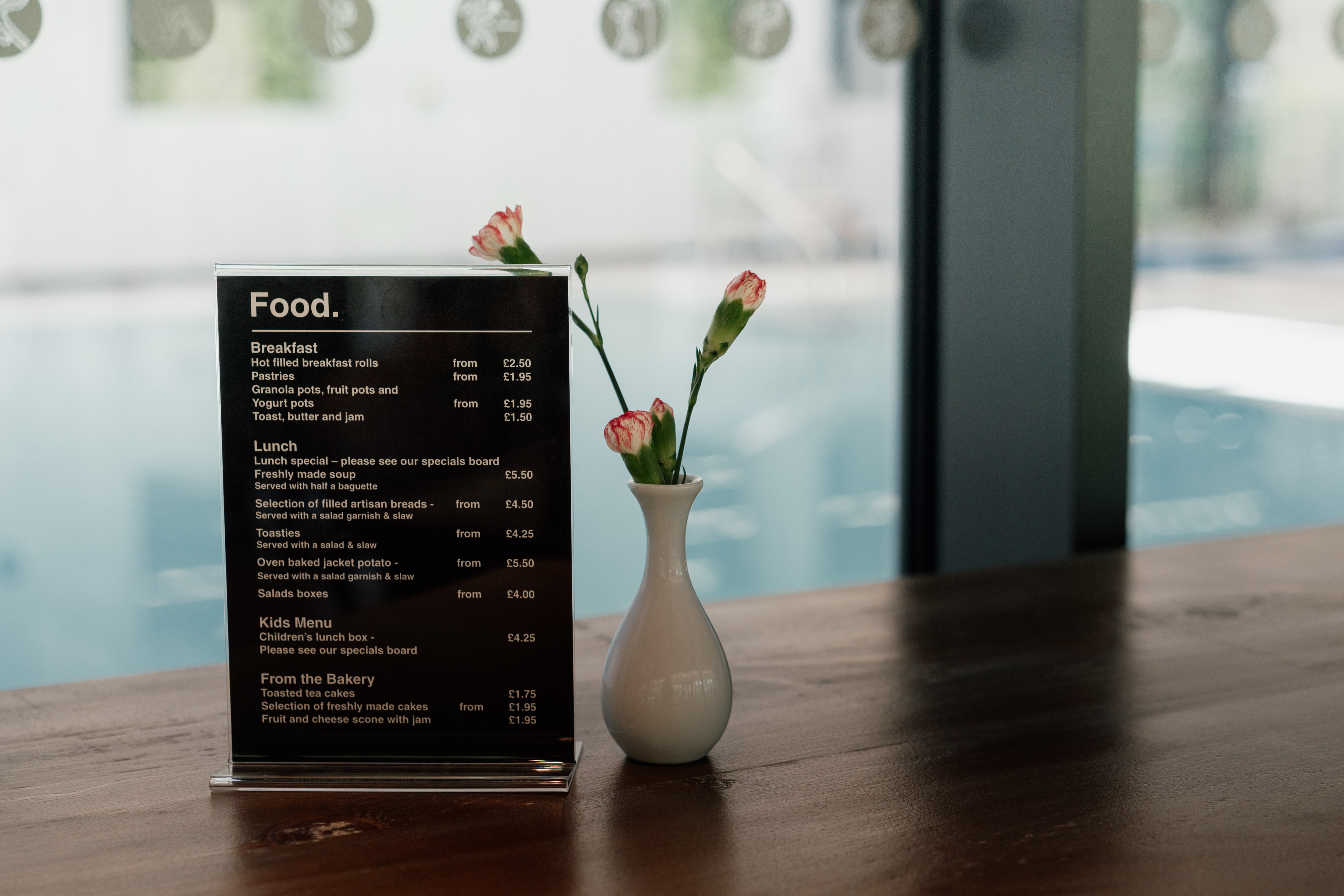 Small food menu sitting on wooden table with small white vase containing carnations, with a view of swimming pool behind