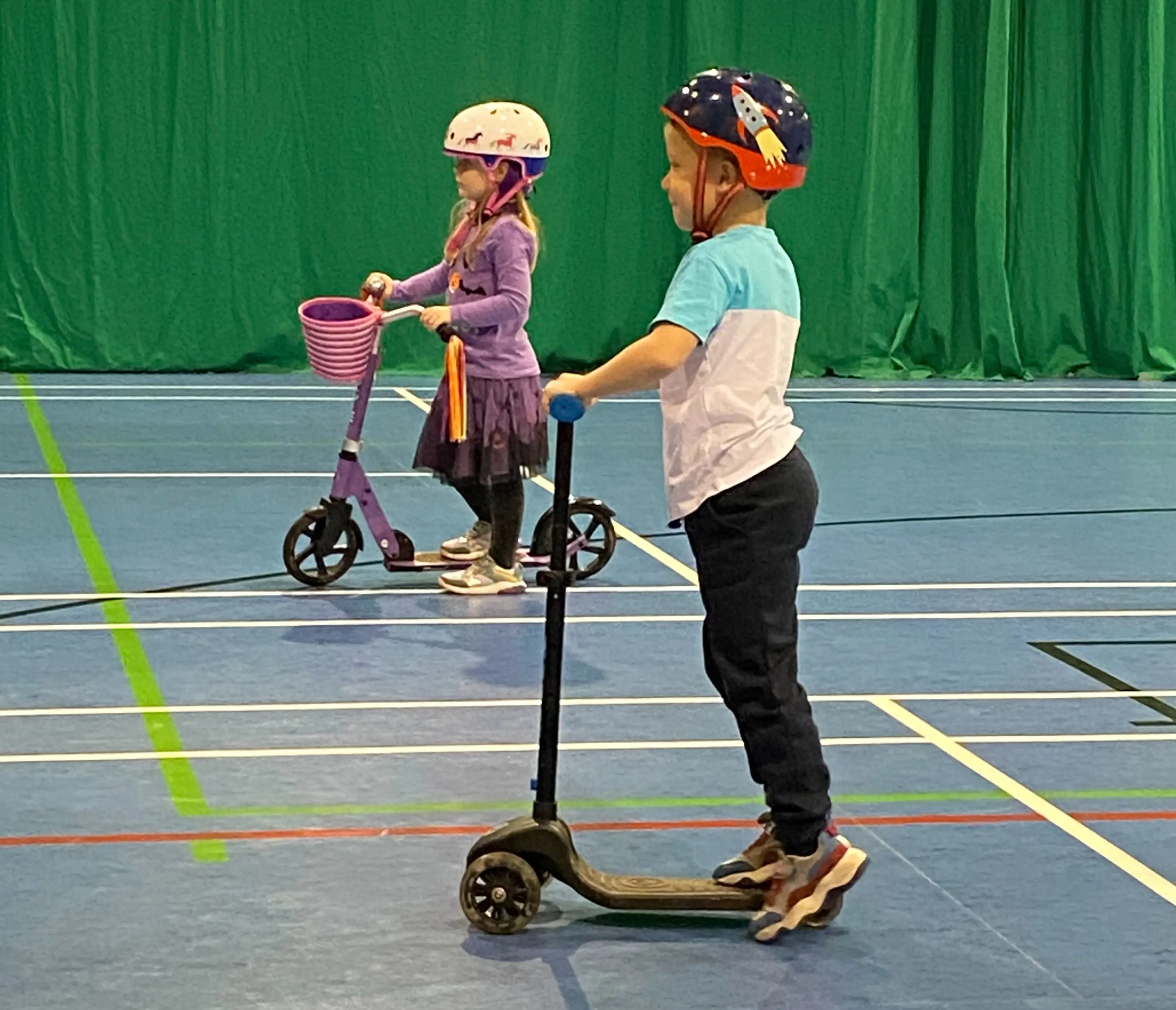 Two children in sports hall wearing helmets and riding scooters