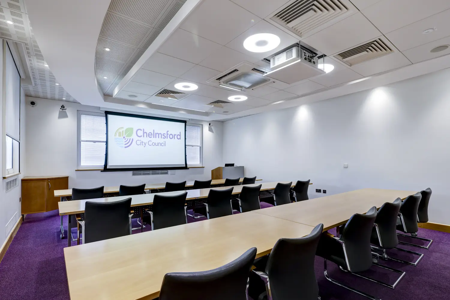 Meeting room set out classroom style, with tables and chairs in rows facing large screen